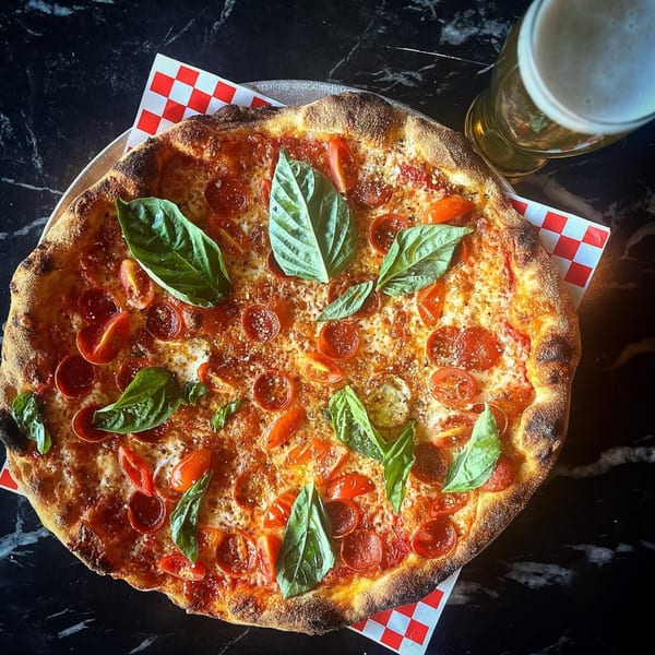 A pepperoni pizza with cherry tomatoes and basil, next to a glass of beer on a dark table.