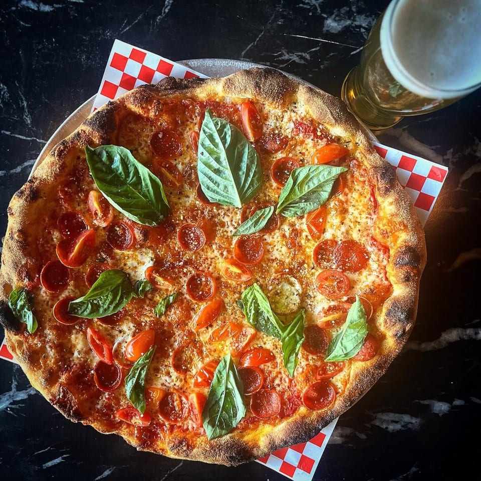 A pepperoni pizza with cherry tomatoes and basil, next to a glass of beer on a dark table.