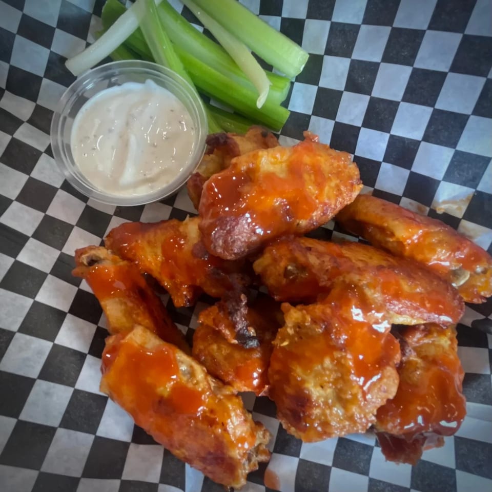Buffalo chicken wings with ranch dressing and celery sticks on checkered paper.